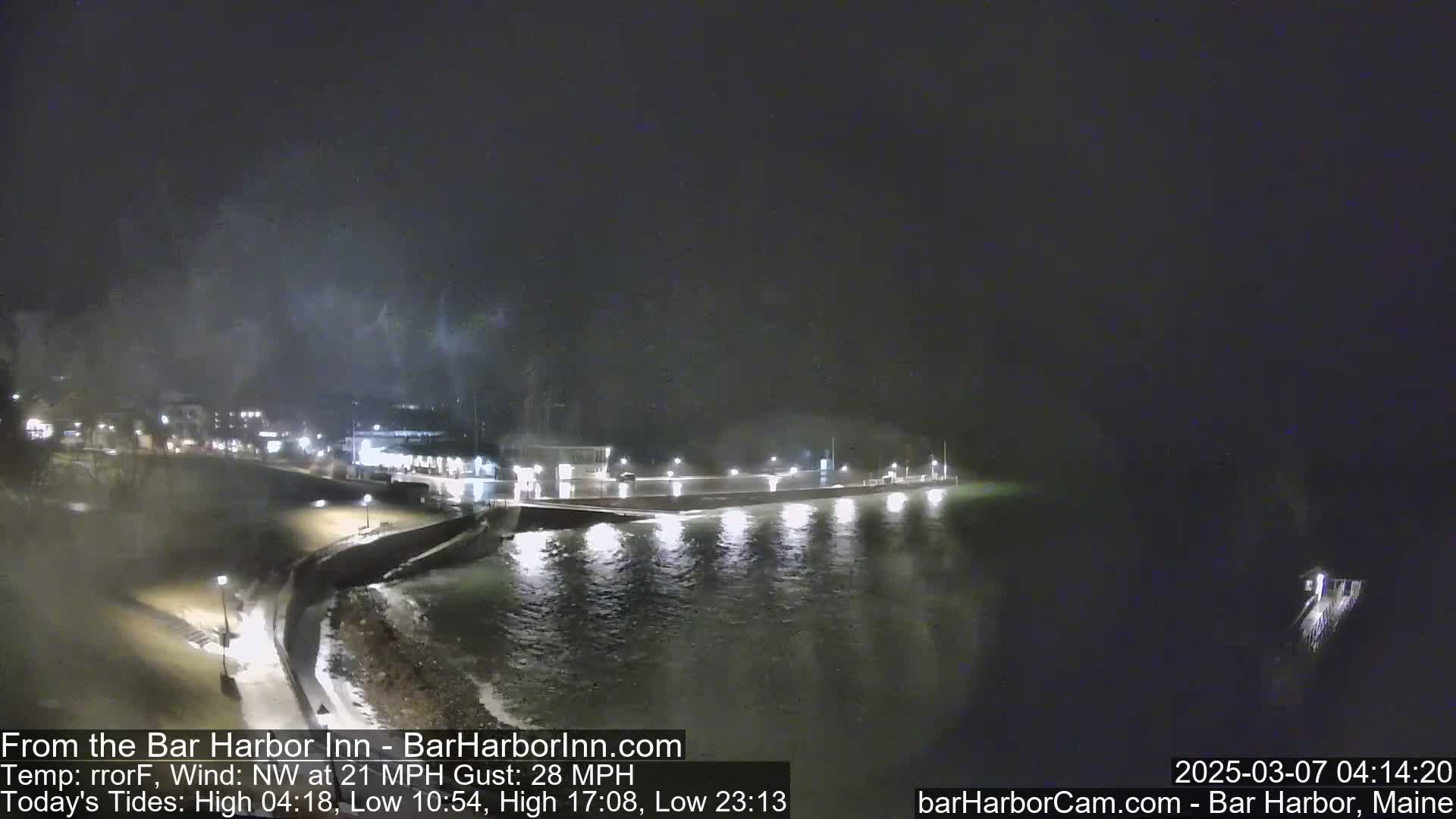A nighttime, coastal scene shows a dark, wavy body of water lapping against a lit seawall, under a cloudy sky with a northwest wind.