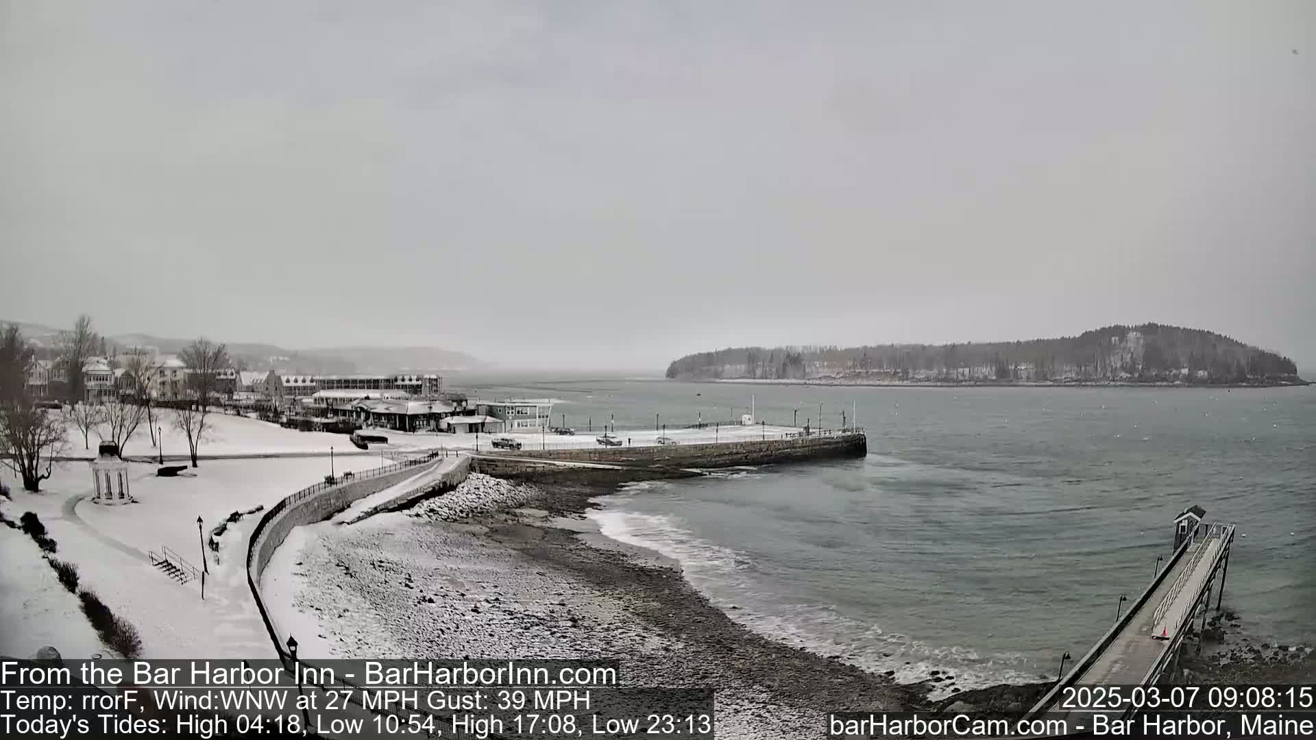 A snow-covered coastal town with buildings, a pier, and a small island in the distance under an overcast sky.