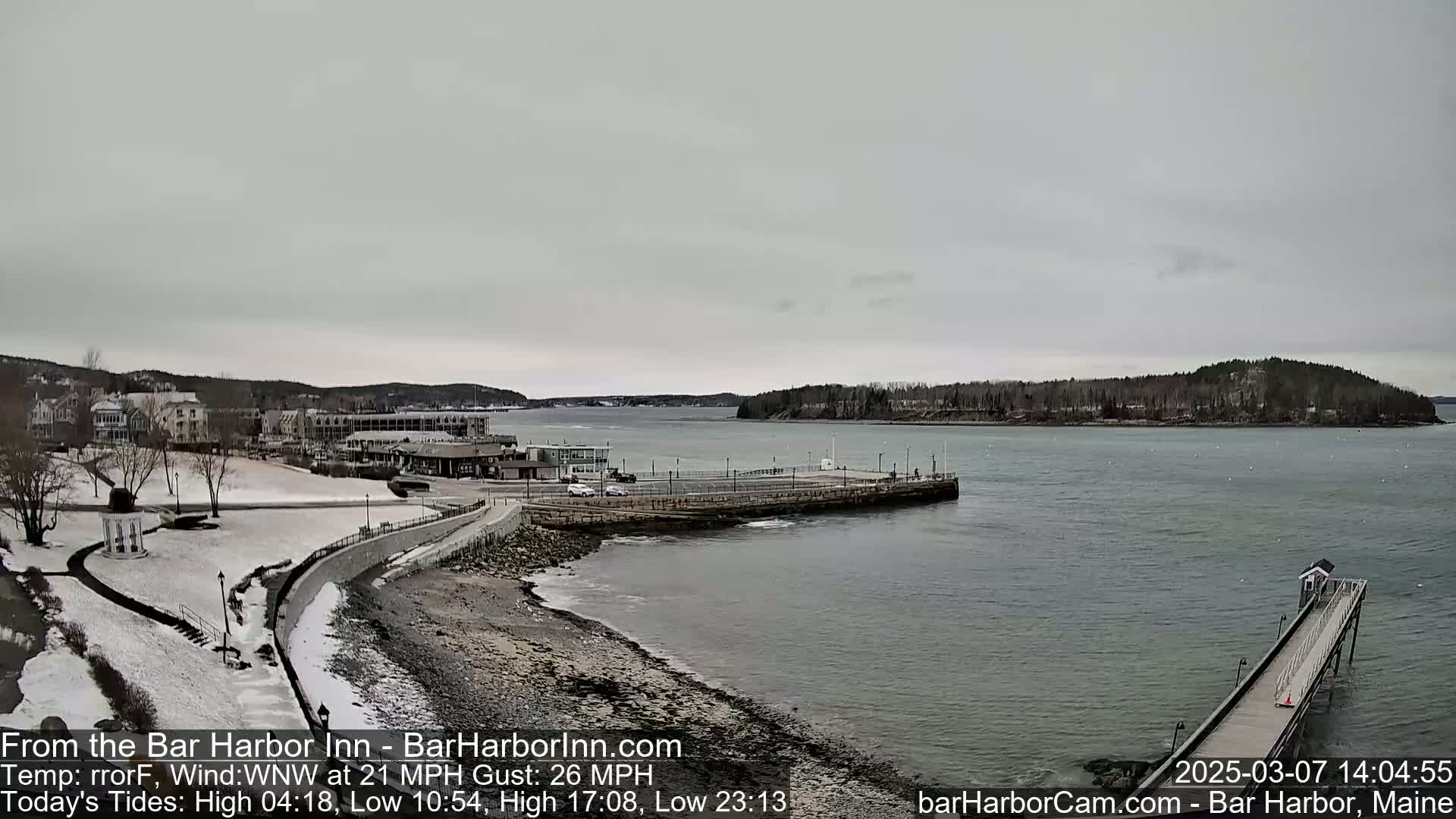 A coastal town with snow-covered ground, a partially frozen bay, and a long pier extending into the water under an overcast sky.
