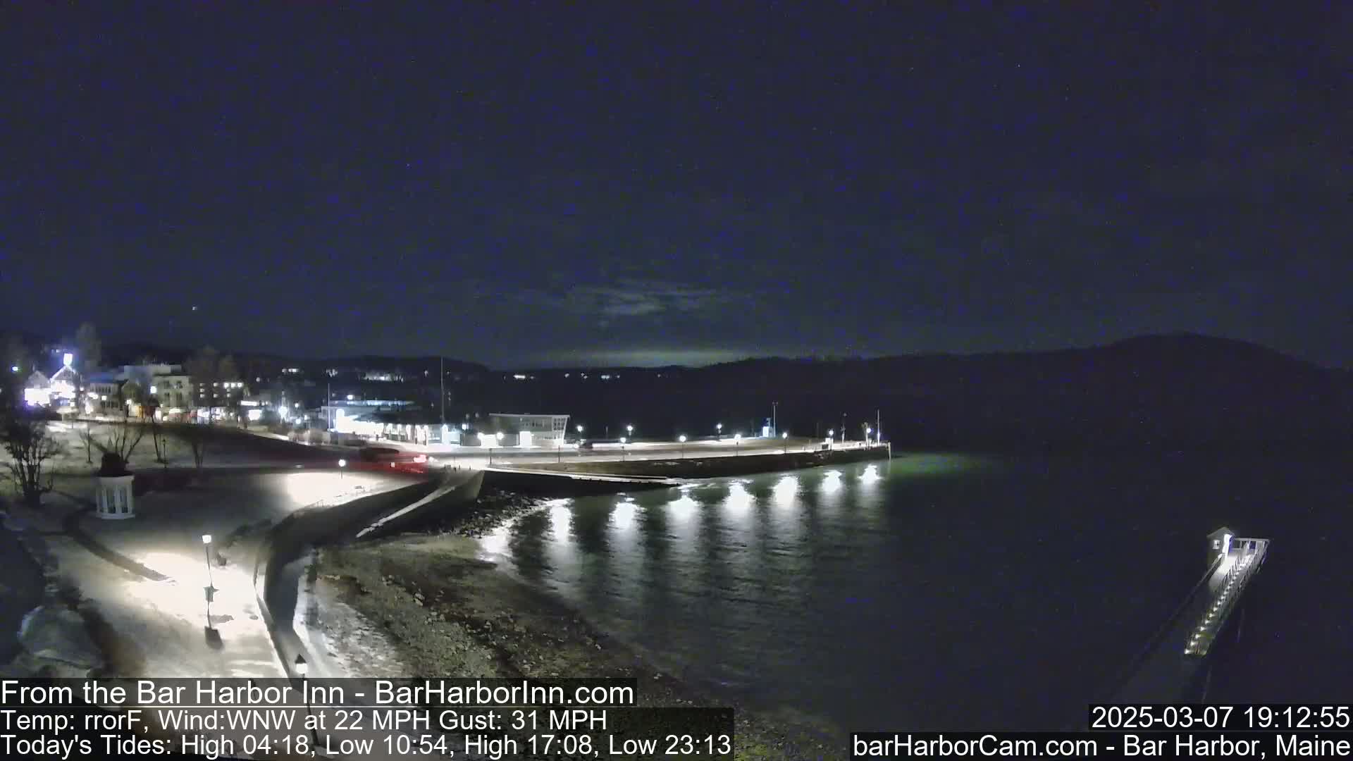 A nighttime coastal scene shows a town's waterfront with a pier extending into calm water under a dark, clear sky with a wind of 22 mph gusting to 31 mph.