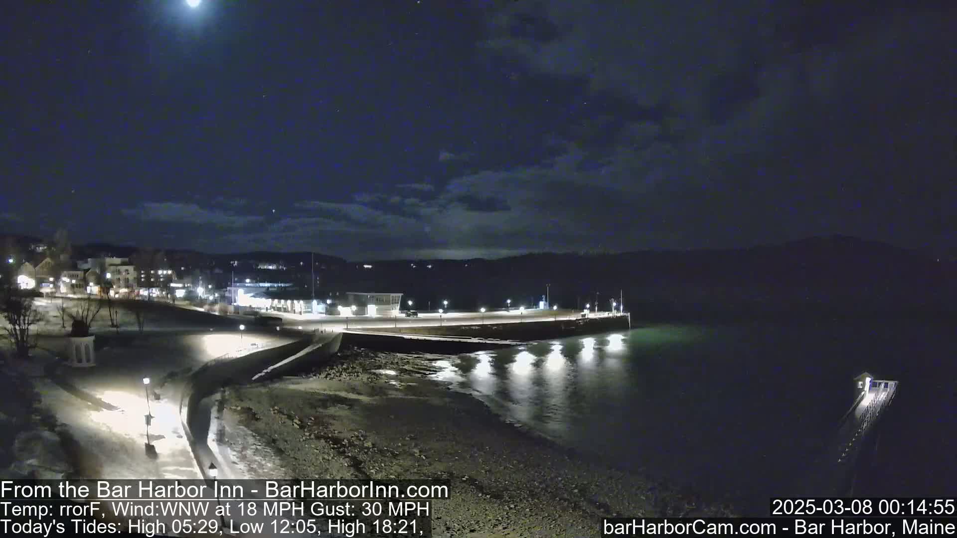 A nighttime view of a town's waterfront, under a mostly clear sky with a bright moon, shows a calm body of water reflecting lights from the shore.