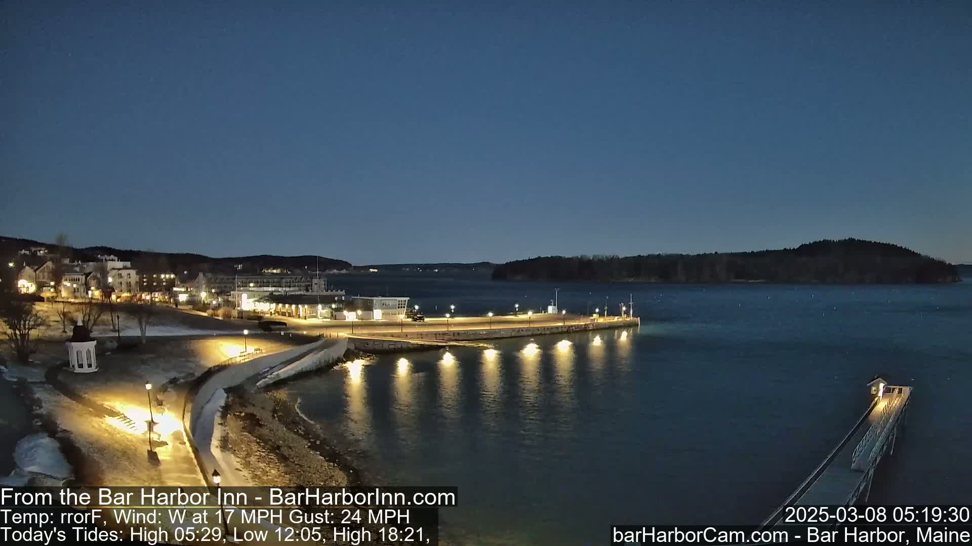 A nighttime, snow-dusted waterfront scene shows a town's illuminated shoreline with a pier extending into calm water under a dark blue sky.