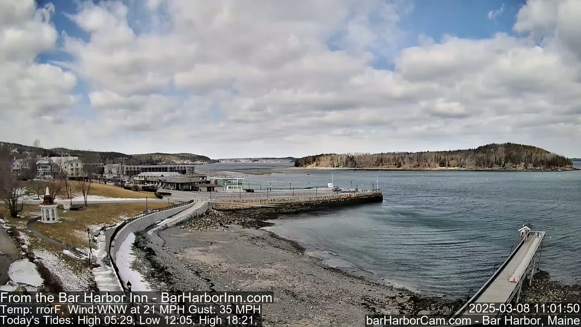 A partly cloudy day reveals a coastal town with a stone walkway leading to a harbor, a small island in the distance, and a pier extending into the water, with some patches of snow visible on the ground.