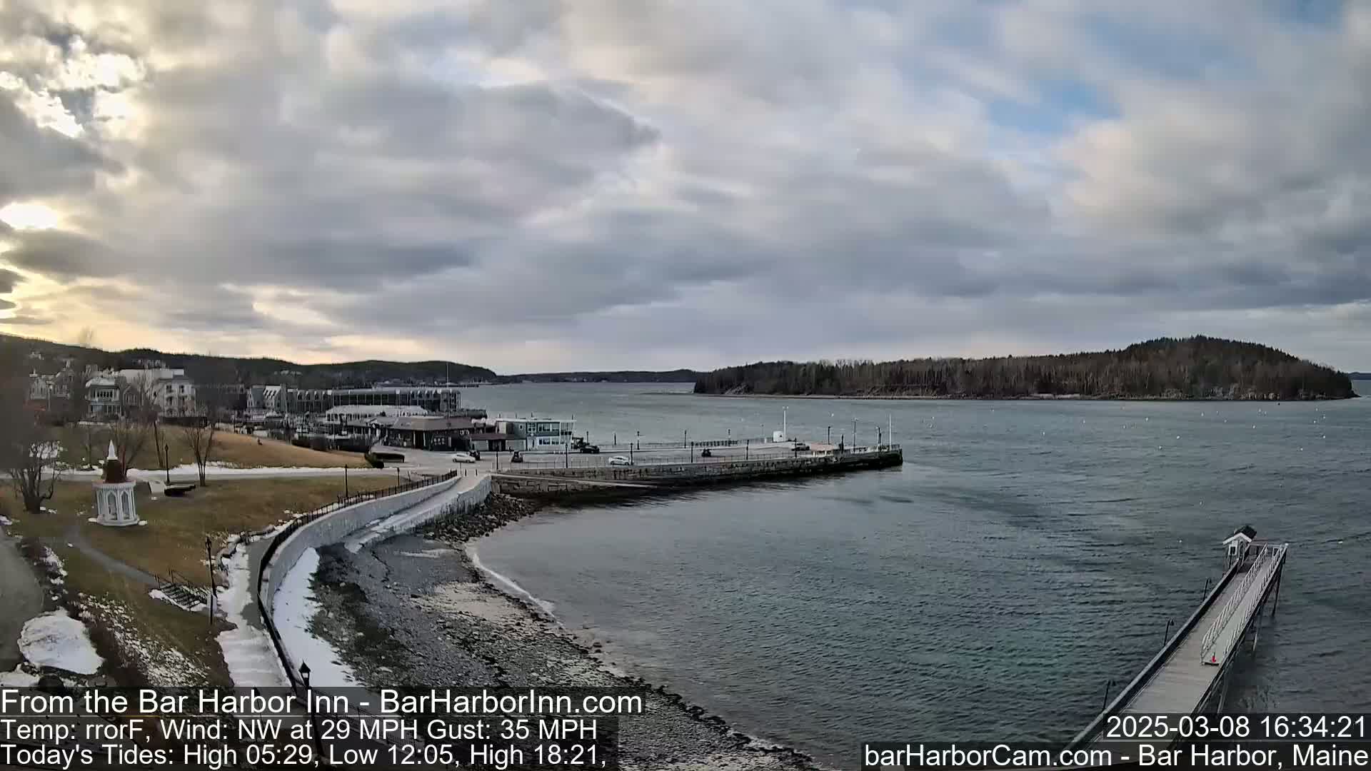 A coastal town with a harbor, a long pier, and a small island in the distance under a cloudy sky.