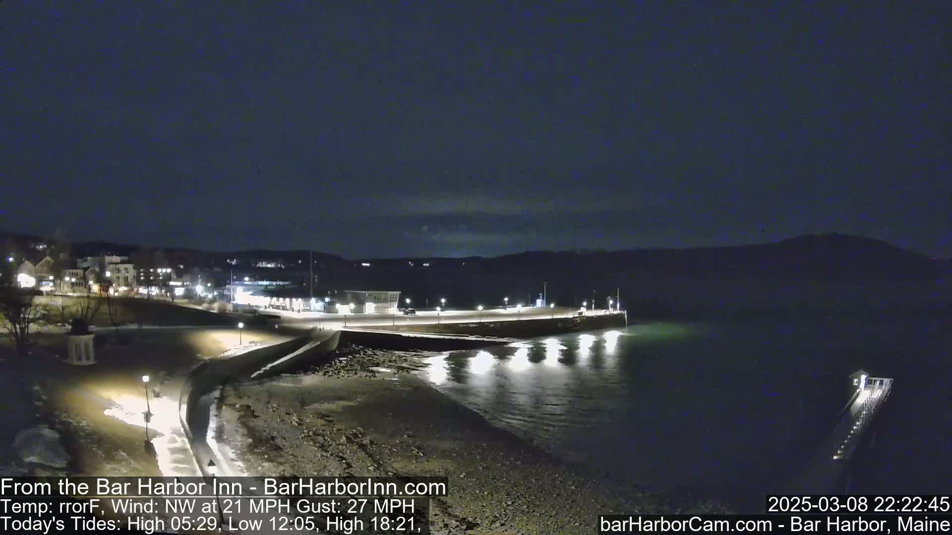 A nighttime, coastal scene shows a dark body of water reflecting lights from a nearby town and waterfront structures under a clear, dark sky.