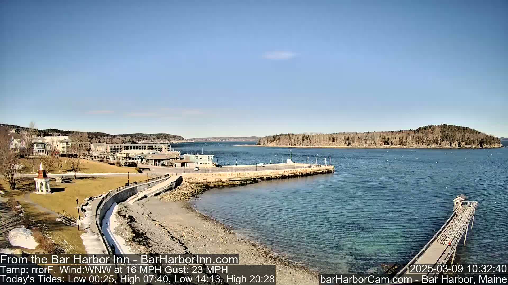 A seaside town with buildings, a grassy area, a stone walkway, a sandy beach, and a long pier extends into a calm, blue body of water under a clear, sunny sky.