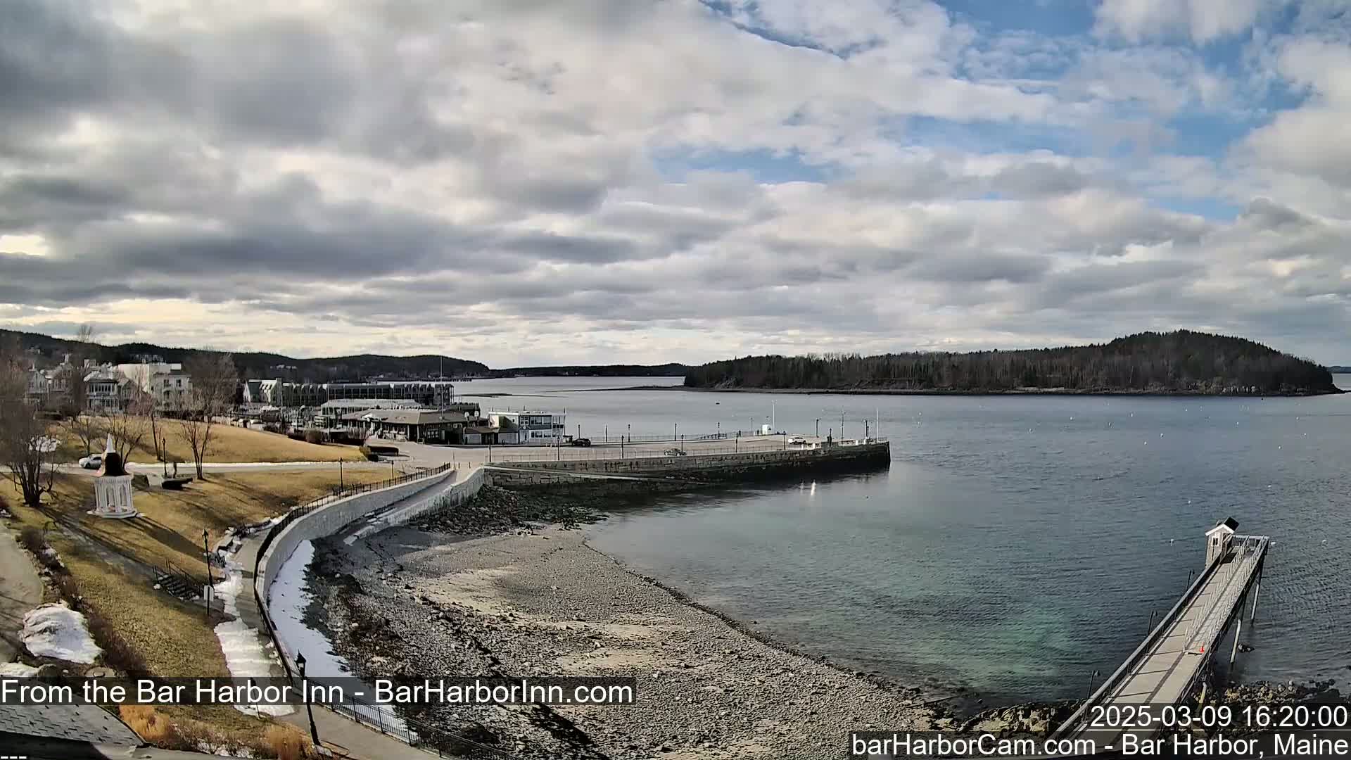 A rocky shoreline with a partially snow-covered grassy area curves around a harbor, with buildings and a small island in the background under a cloudy sky.