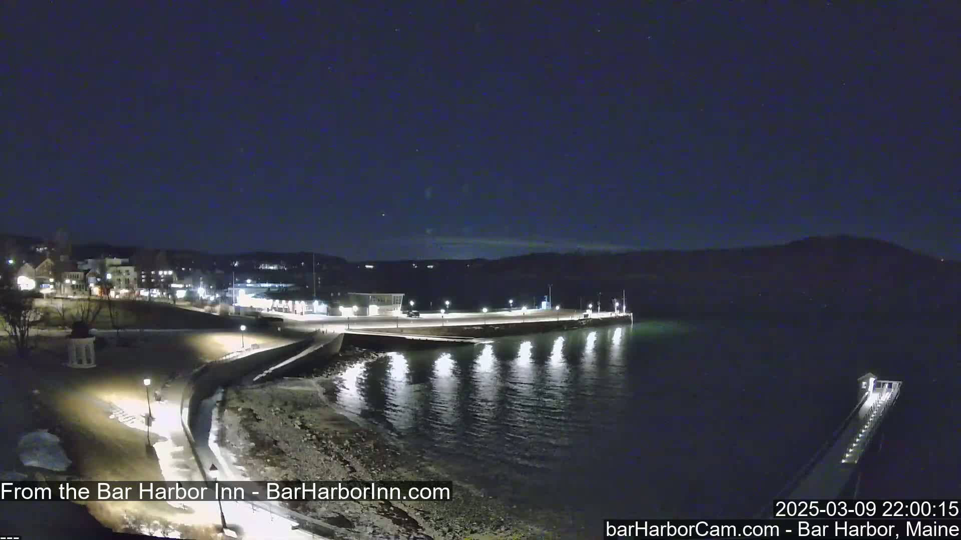 A nighttime waterfront scene shows a town illuminated against a dark, clear sky, with a calm body of water reflecting lights.