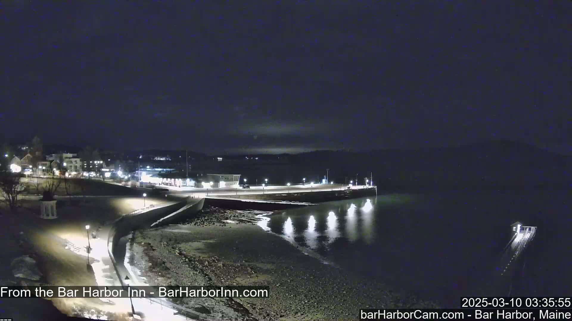 A nighttime coastal scene shows a dark, calm body of water reflecting lights from a shoreline with buildings and a pier.
