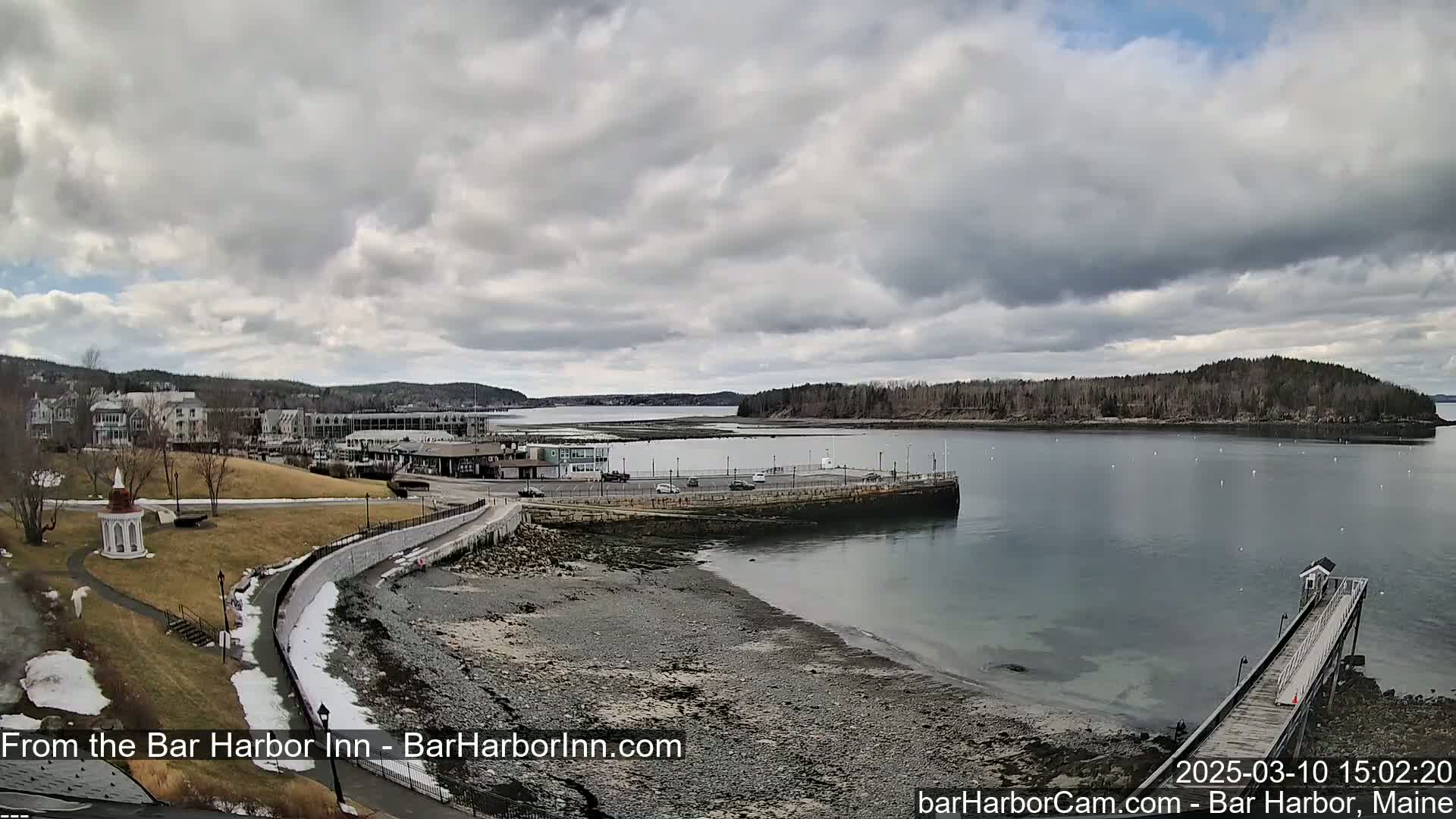 A coastal town with buildings and a grassy area is partially covered in snow under a cloudy sky, next to a calm body of water with a pier extending into it.