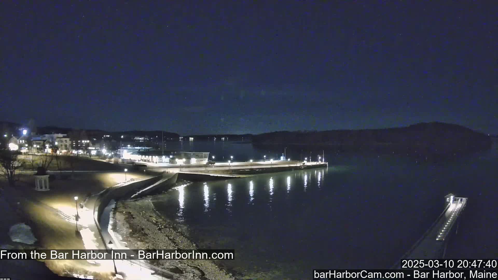 A nighttime coastal scene shows a town's waterfront with lights illuminating a paved walkway, a pier extending into calm dark water, and a small amount of snow on the ground.