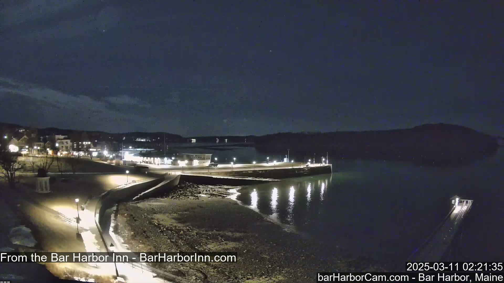 A nighttime view of a harbor town's waterfront, with a calm body of water reflecting lights from the shore under a dark, mostly clear sky.