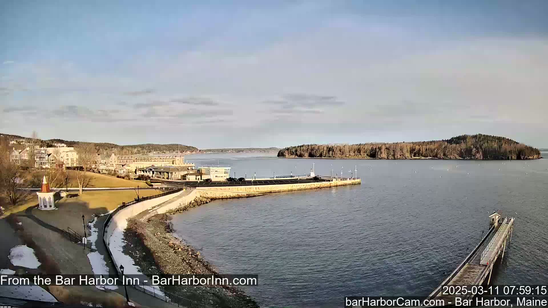 A partly sunny day reveals a coastal town with buildings, a harbor, a long pier, and a small island in the distance, with some snow visible on the ground near the water.