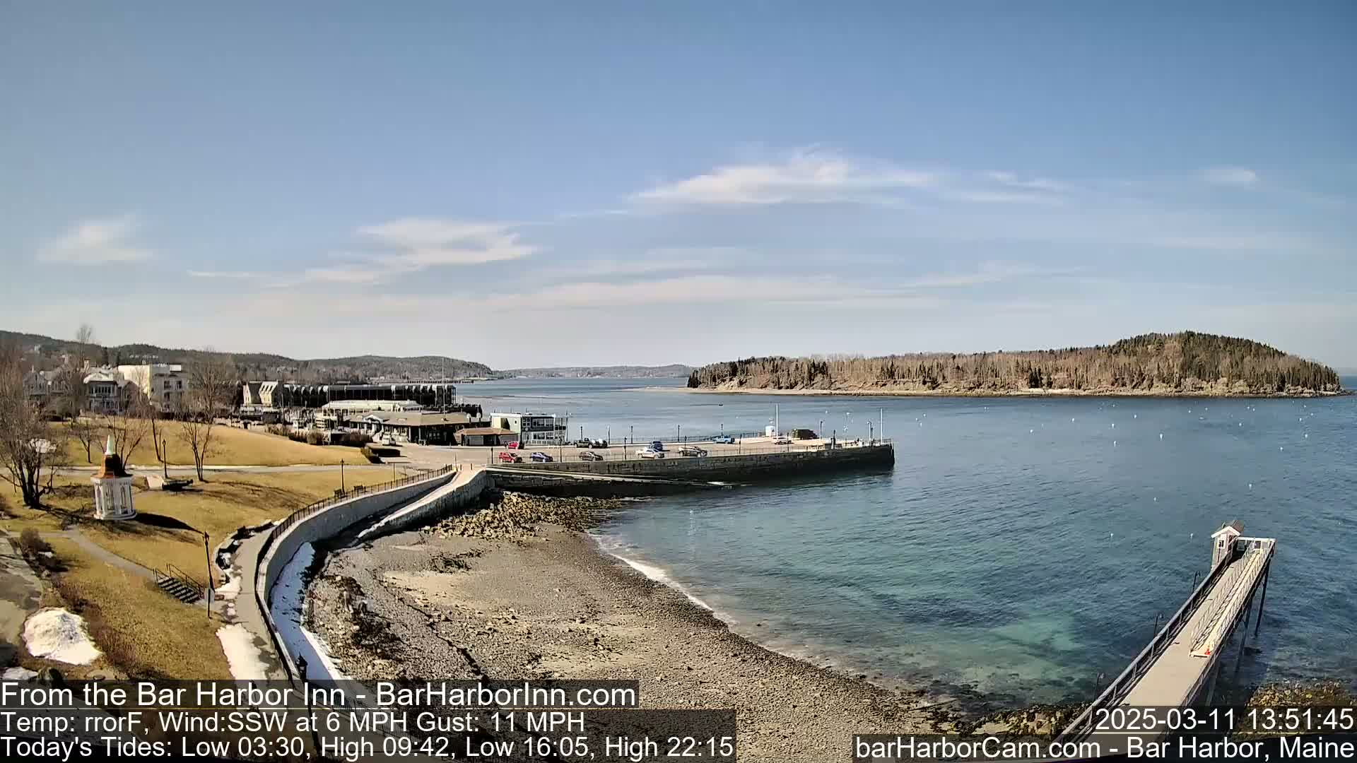 A mostly sunny day reveals a rocky shoreline, a calm harbor with a small island in the distance, and a town with buildings and a parking lot visible in the background.
