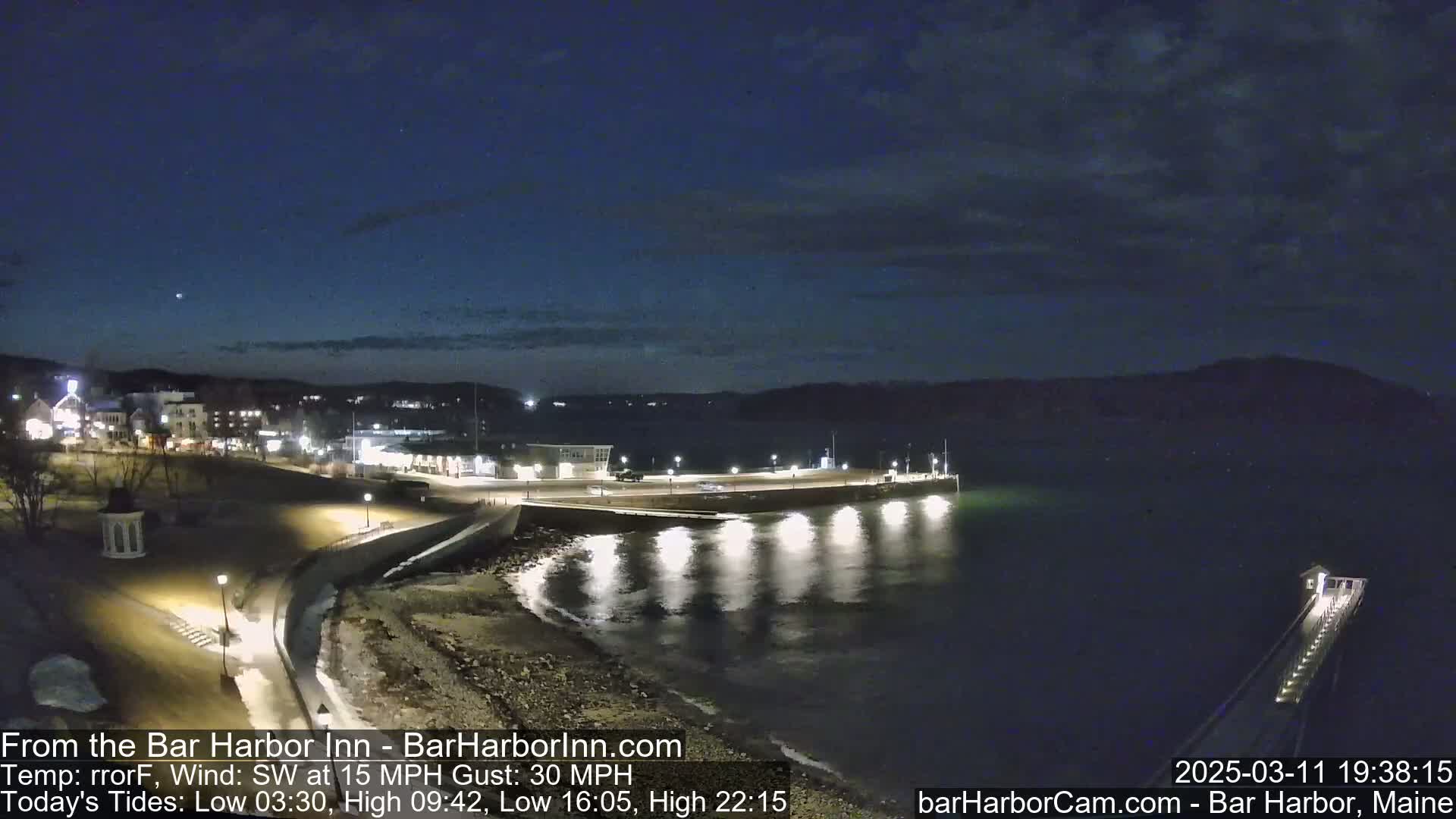 A nighttime view of a harbor town with a pier extending into dark water under a partly cloudy sky, with a 15 mph southwest wind and gusts up to 30 mph.