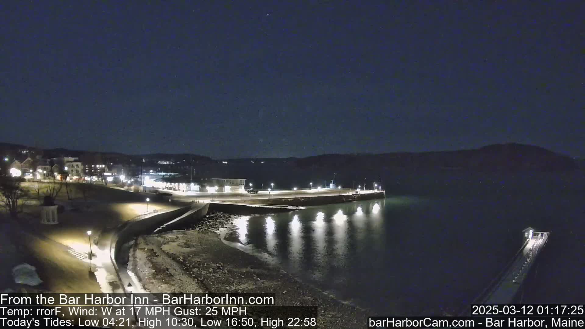 A nighttime view of a waterfront town with a calm body of water reflecting lights from the shore, under a clear, dark sky.