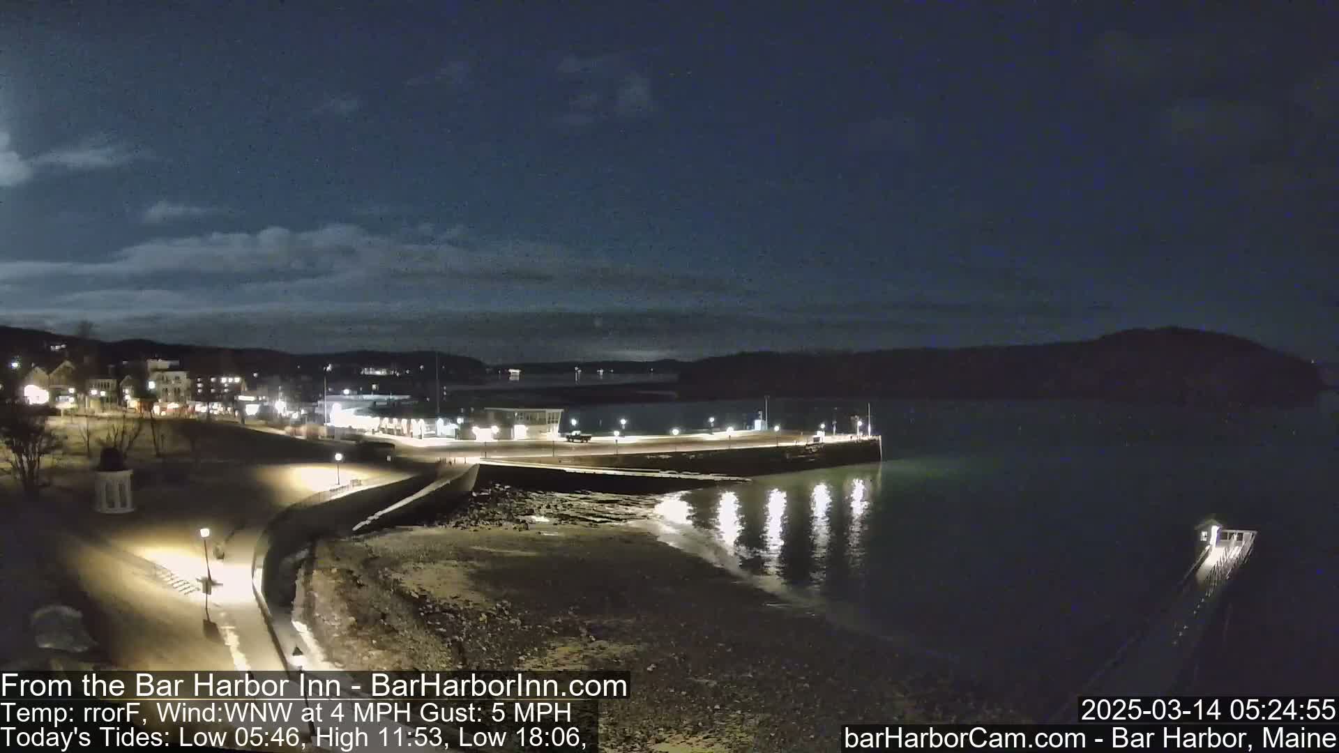 A nighttime view of a harbor town's waterfront, with a calm, dark body of water reflecting lights from the shore under a mostly clear, dark sky.