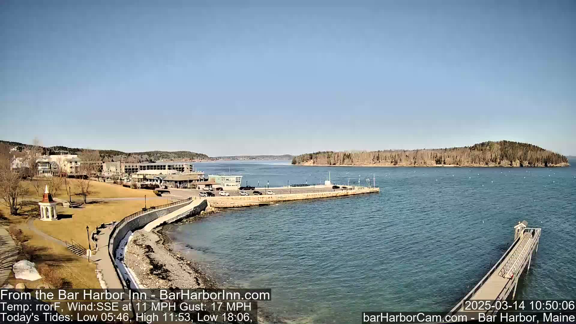 A coastal town scene on a sunny day with a calm body of water, a paved walkway along the shoreline, and a small island in the distance.