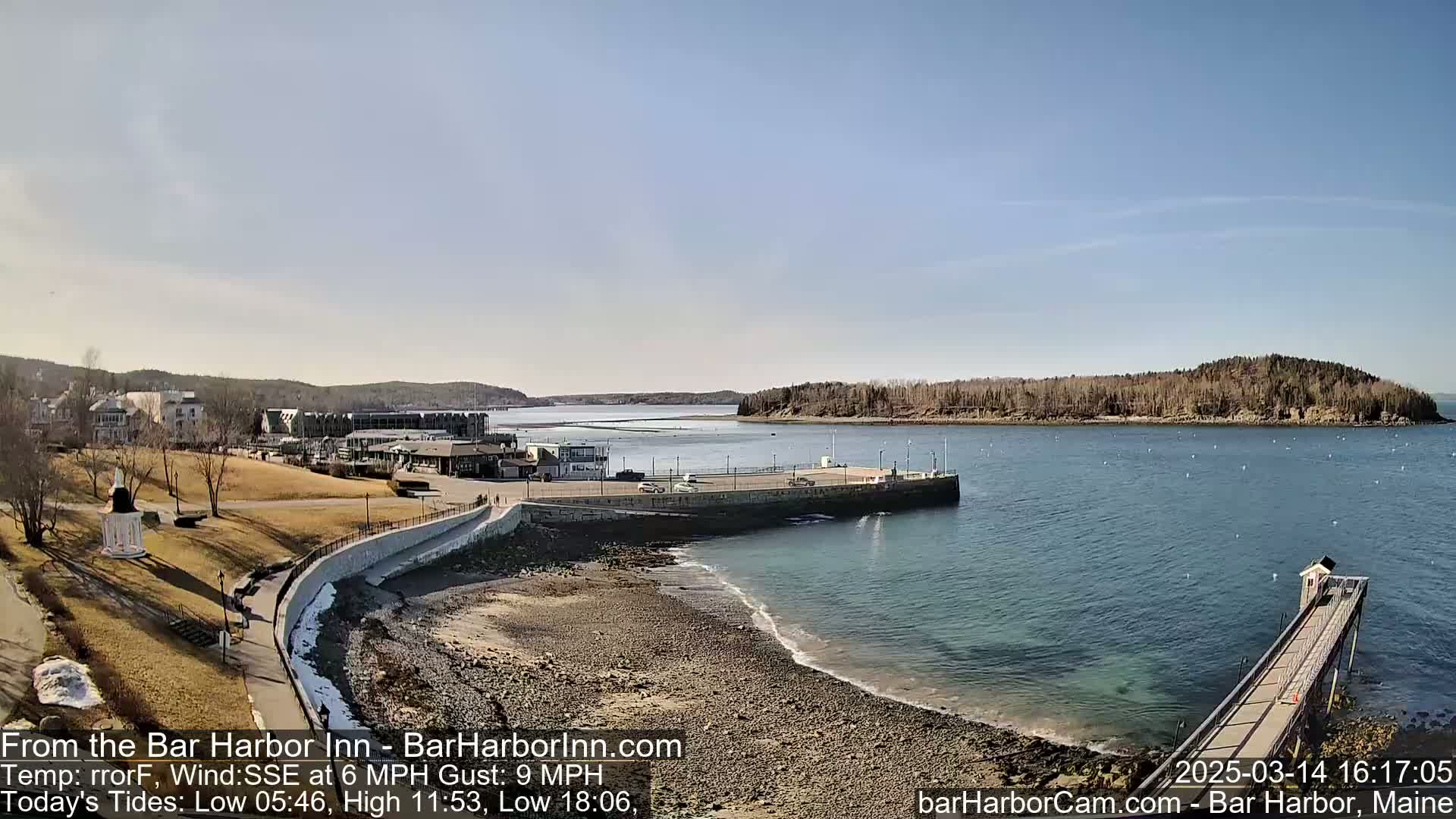 A harbor scene on a sunny day with a calm sea, showing a rocky shore, a pier extending into the water, and buildings in the background.