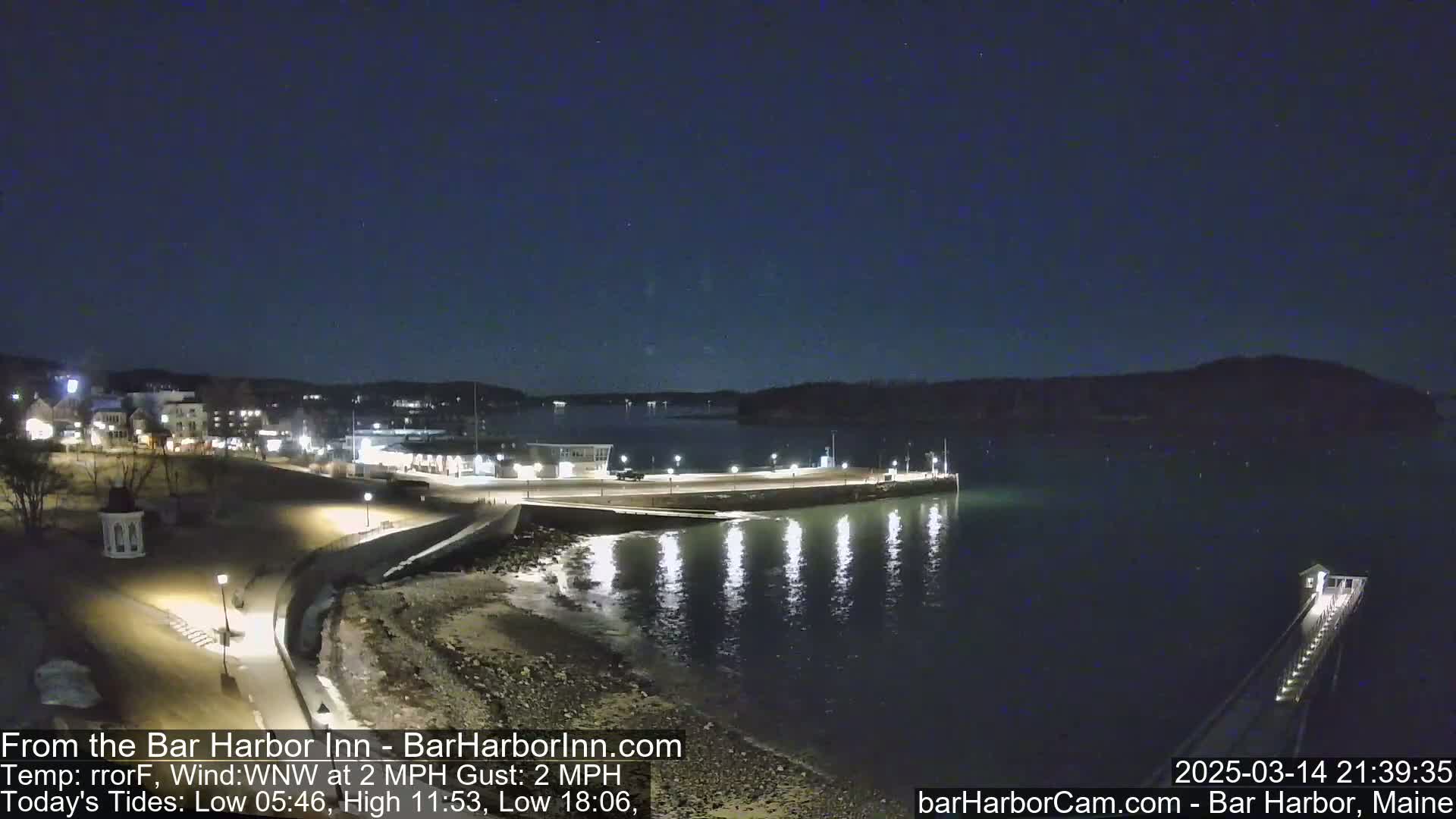 A nighttime view of a harbor with lights illuminating a waterfront area and a pier extending into the calm dark water under a clear, starry sky.