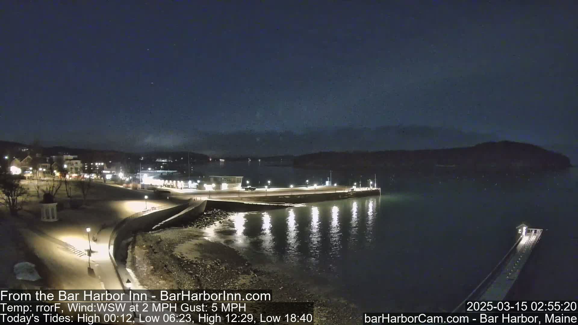 A nighttime view of a town's waterfront, showing a calm, dark body of water with lights reflecting on its surface, under a clear, starry sky.