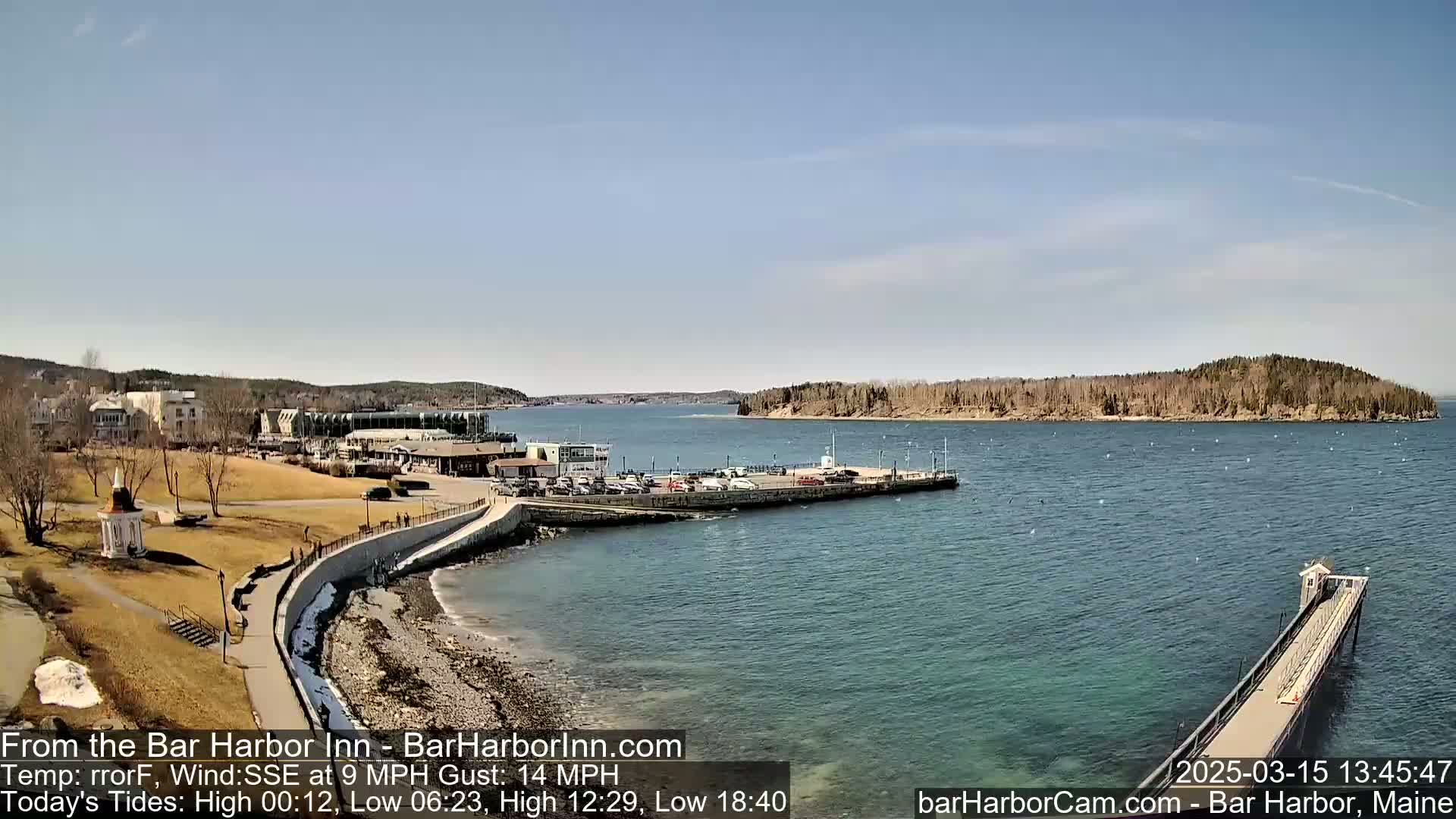 A clear, sunny day shows a coastal town with a harbor, a long pier extending into the water, and a small island in the distance.