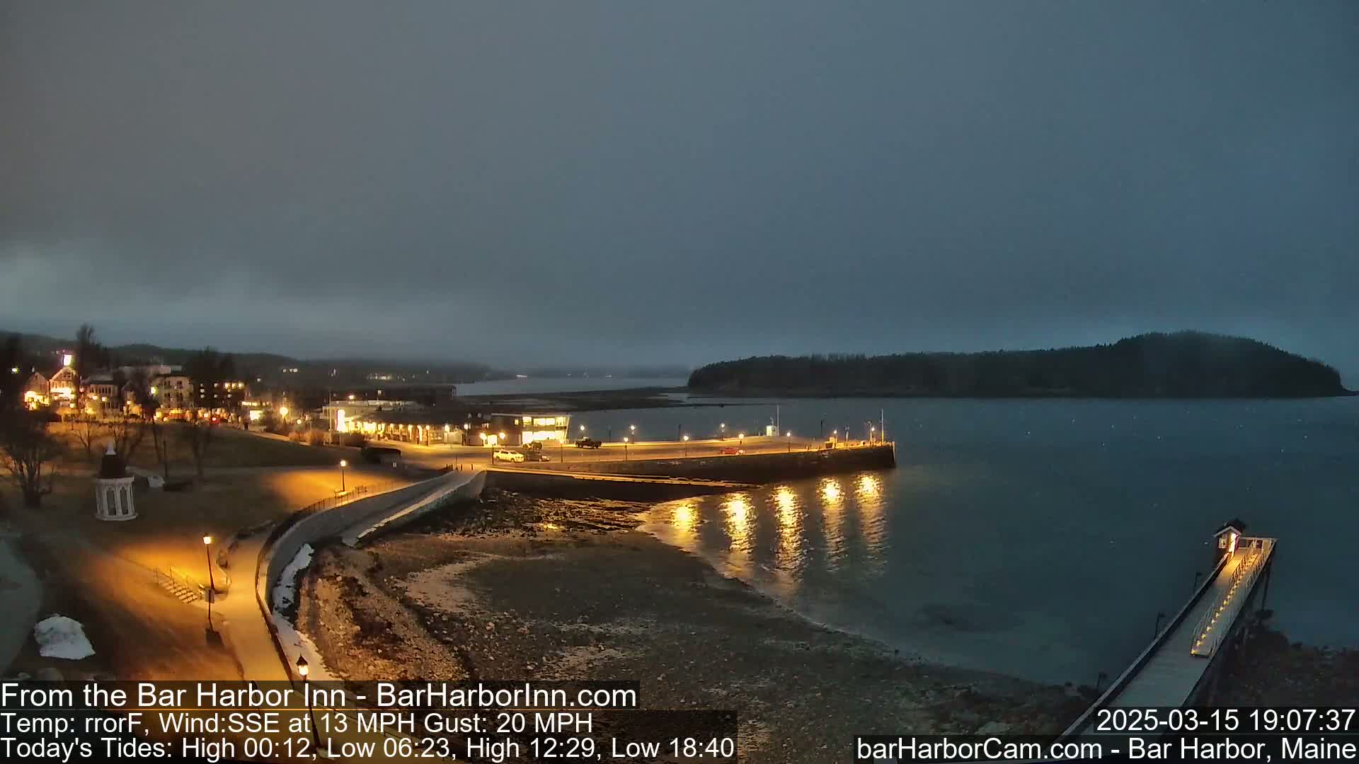 A nighttime coastal scene shows a town with illuminated buildings, a harbor with a pier and lights, and a dark, calm body of water under an overcast sky.