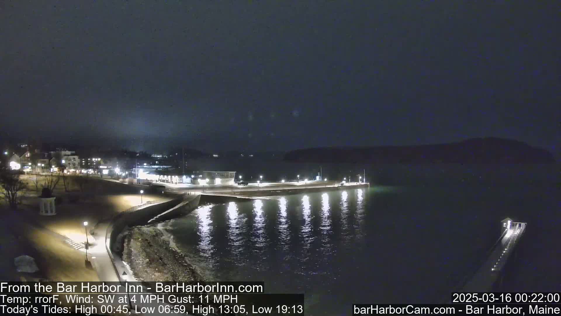 A nighttime coastal scene shows a dark, calm body of water reflecting lights from a pier and buildings along a shoreline under a mostly clear sky.