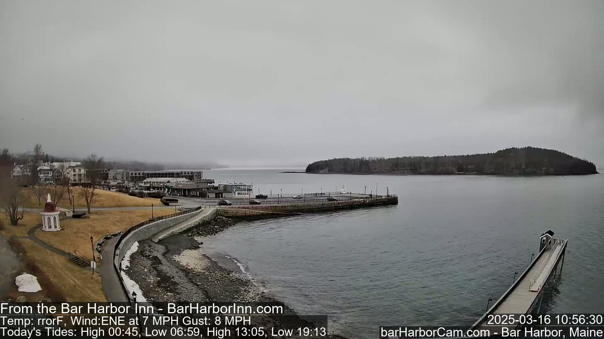 A gray, overcast day shows a harbor town with a stone walkway leading to a pier extending into calm water, with a small island in the distance.