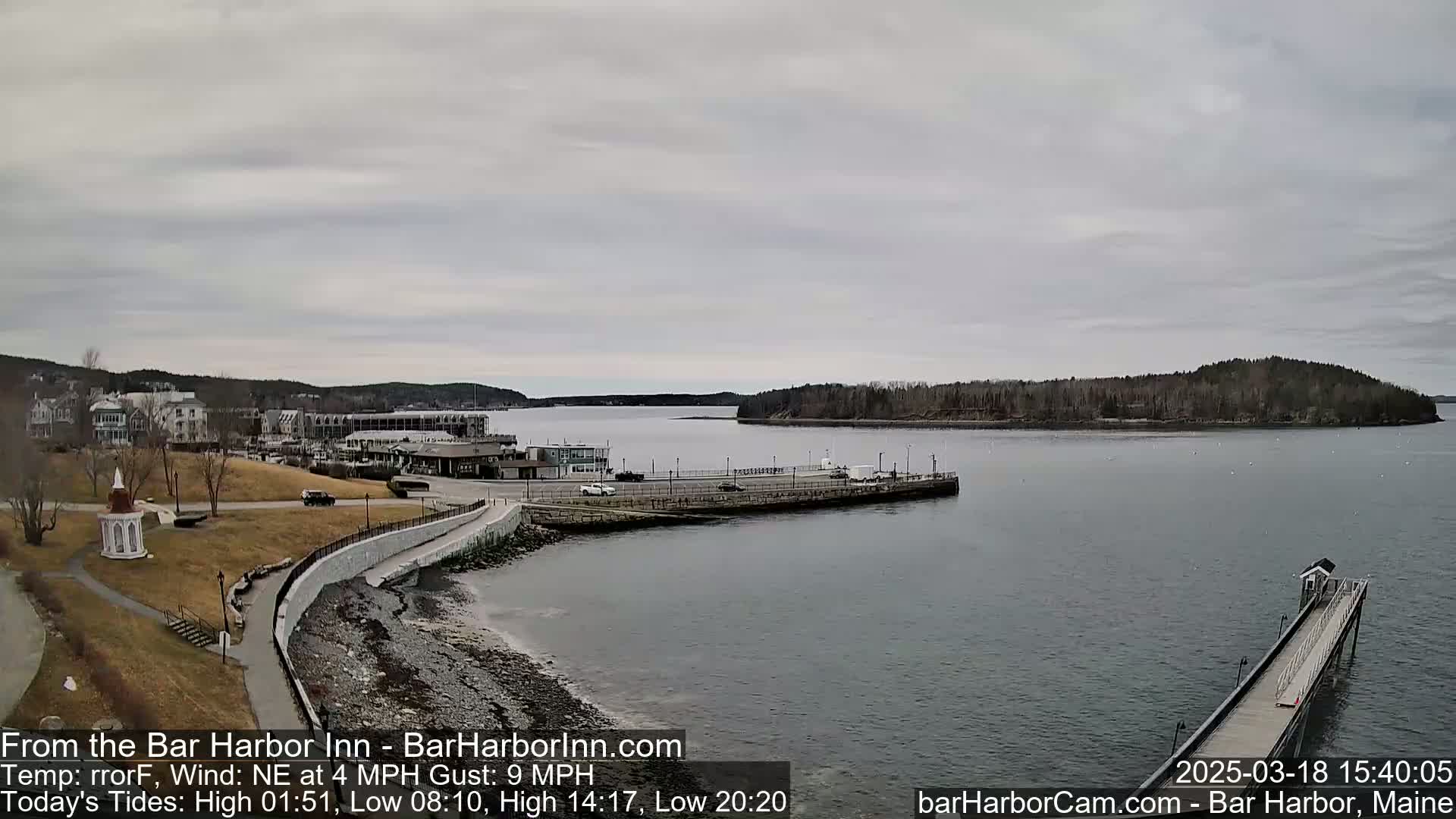 A waterfront town with a calm harbor under an overcast sky is viewed from a slightly elevated position, showing a long pier extending into the water.