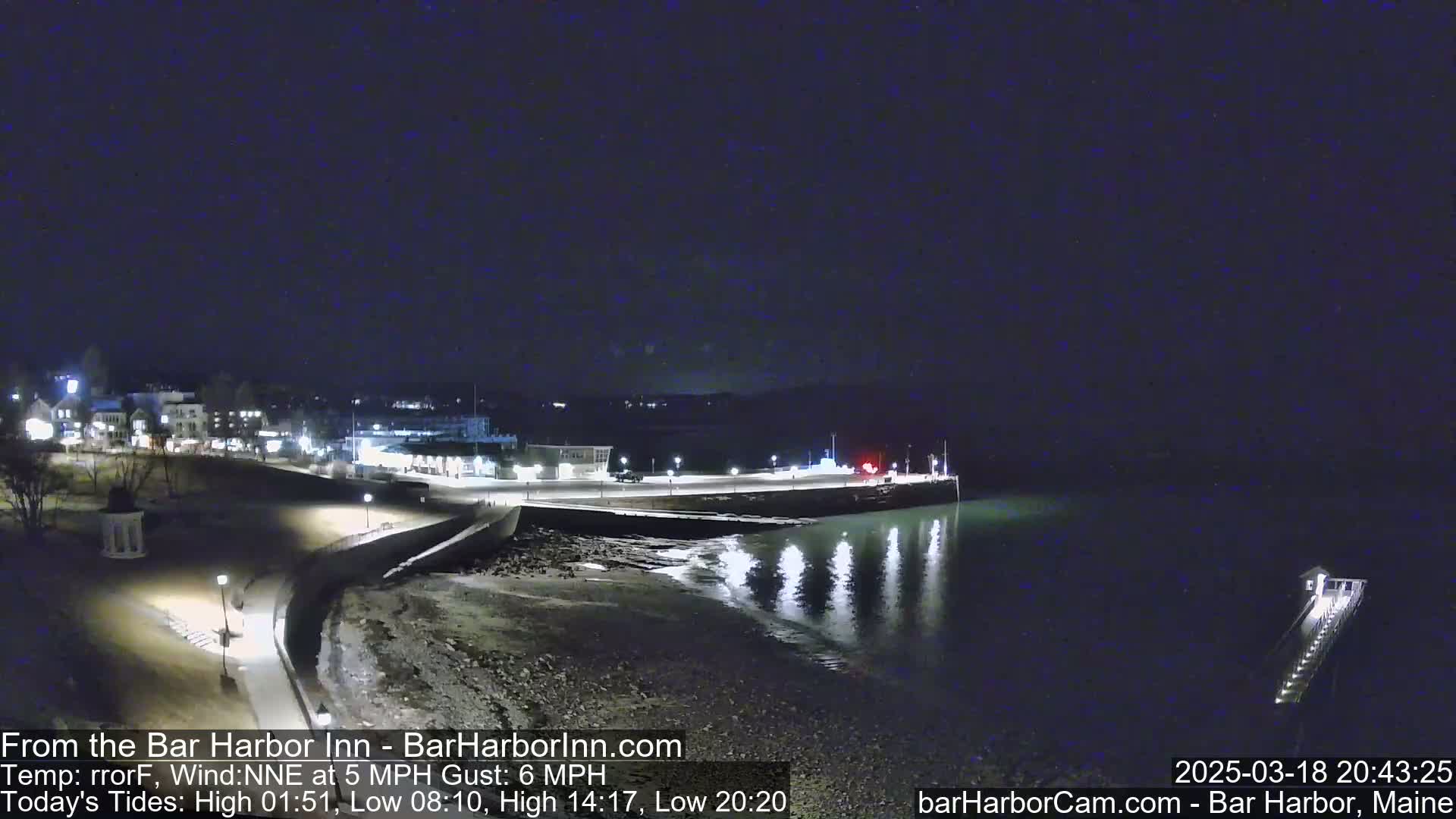 A nighttime view of a calm harbor with a town's lights reflecting on the water, under a dark, clear sky.