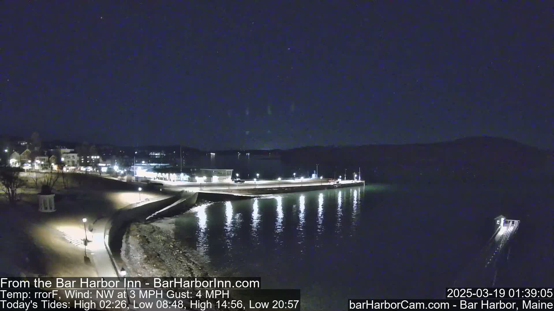 A nighttime coastal scene shows a dark body of water reflecting lights from a town and pier on the shore under a clear, dark sky.