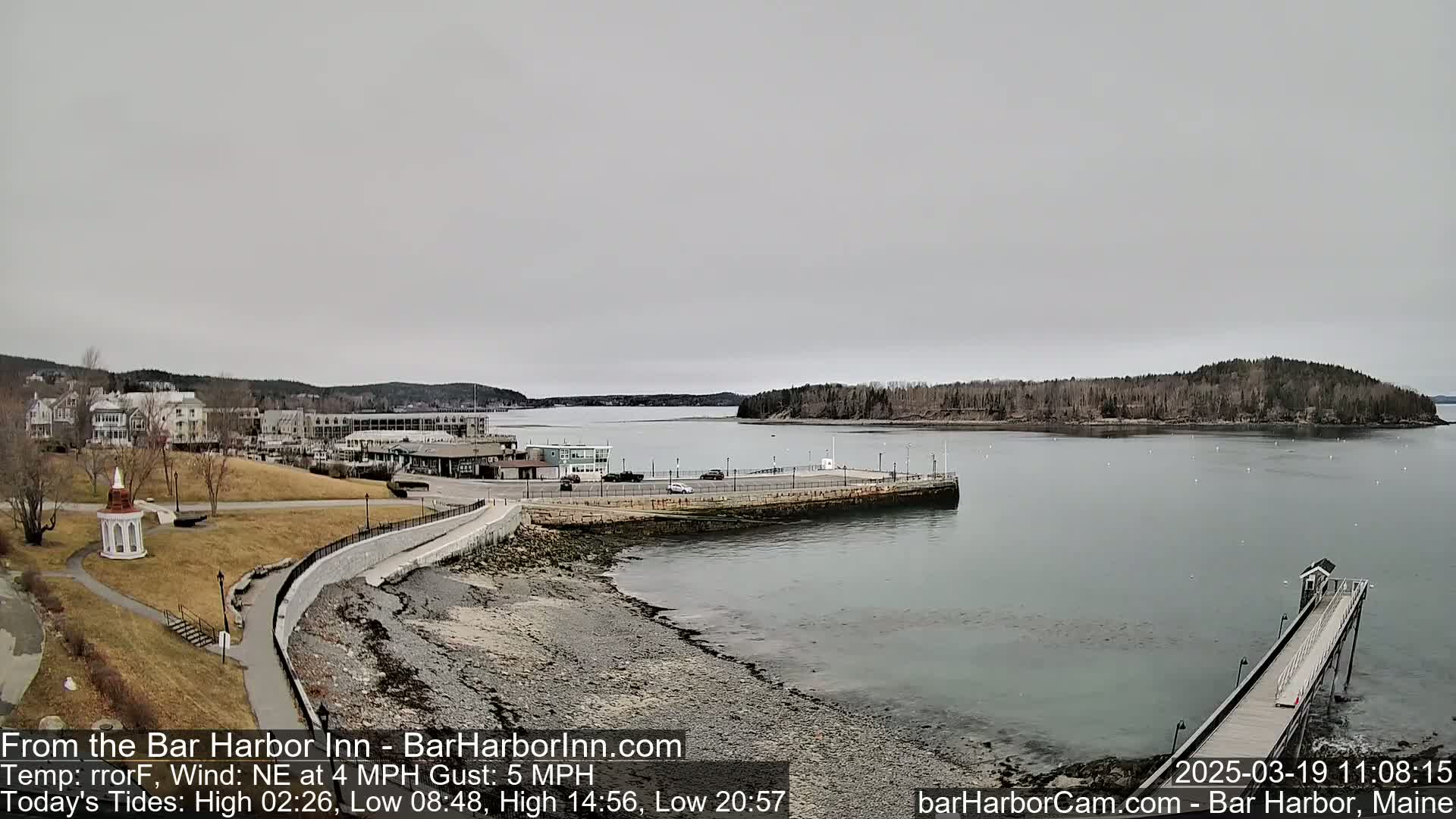 A rocky shoreline with a paved walkway leads to a harbor featuring a pier and buildings under an overcast sky.