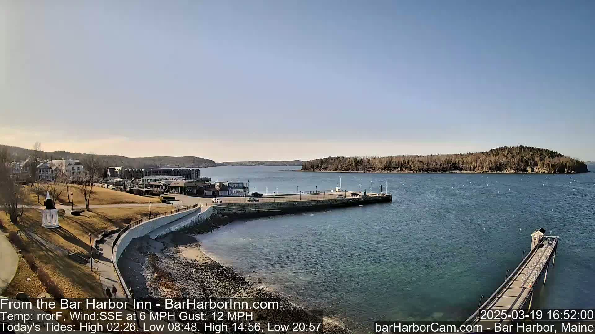 A sunny day reveals a harbor scene with a town, grassy areas, a long pier extending into calm blue water, and a small island in the distance.