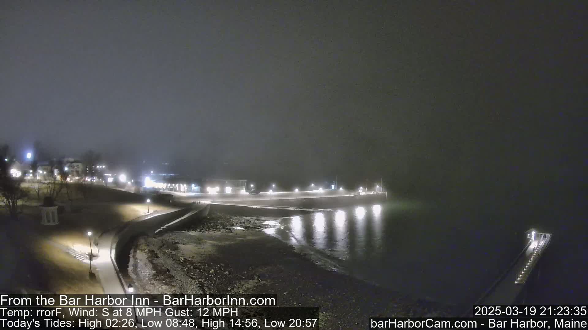A foggy night scene shows a dark, calm body of water reflecting lights from a shoreline with buildings and a pier.