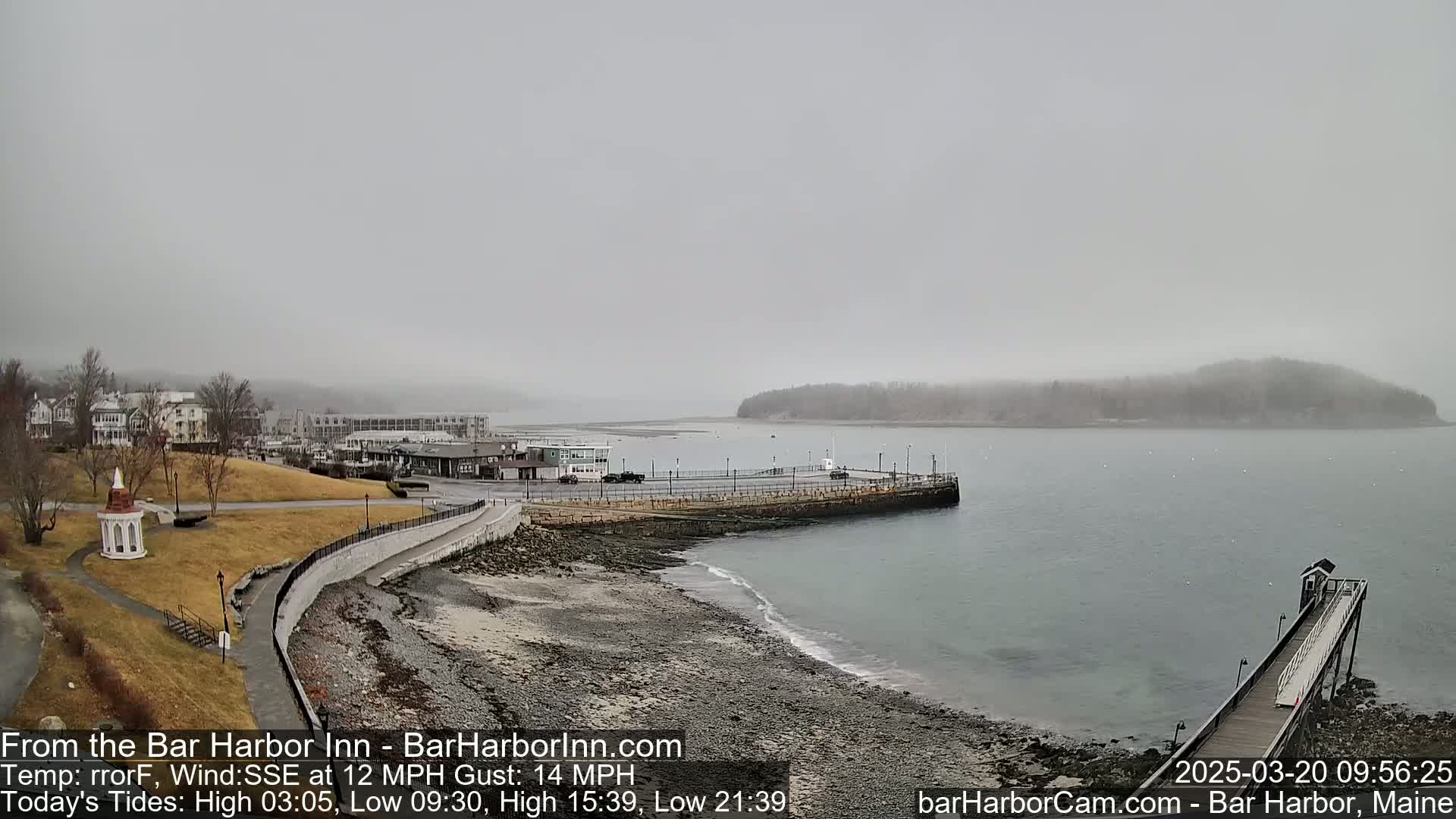 A foggy seaside town is seen from an elevated vantage point, showing a rocky shoreline, a long pier, and a calm body of water with a small island in the distance.