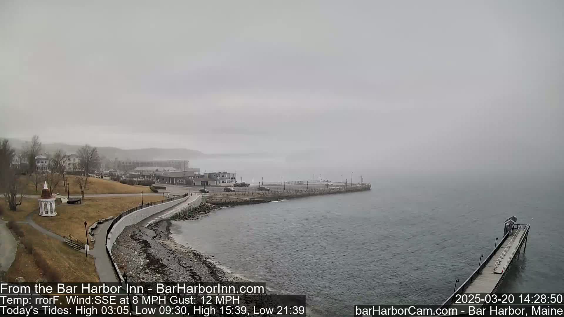 A foggy seaside town is seen from a high vantage point, showing a coastal path, buildings, and a pier extending into the calm water.