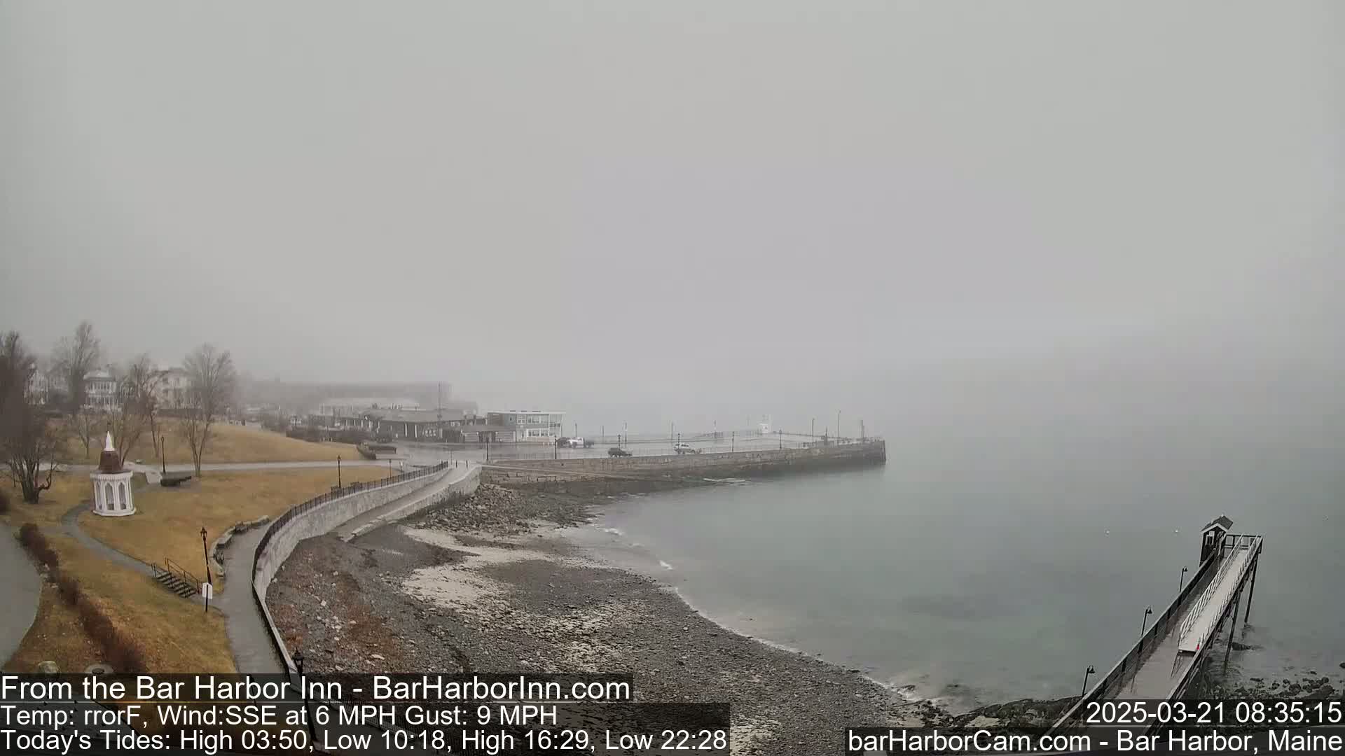 A foggy, overcast coastal scene shows a rocky shoreline with a paved walkway, buildings in the distance, and a long pier extending into the water.