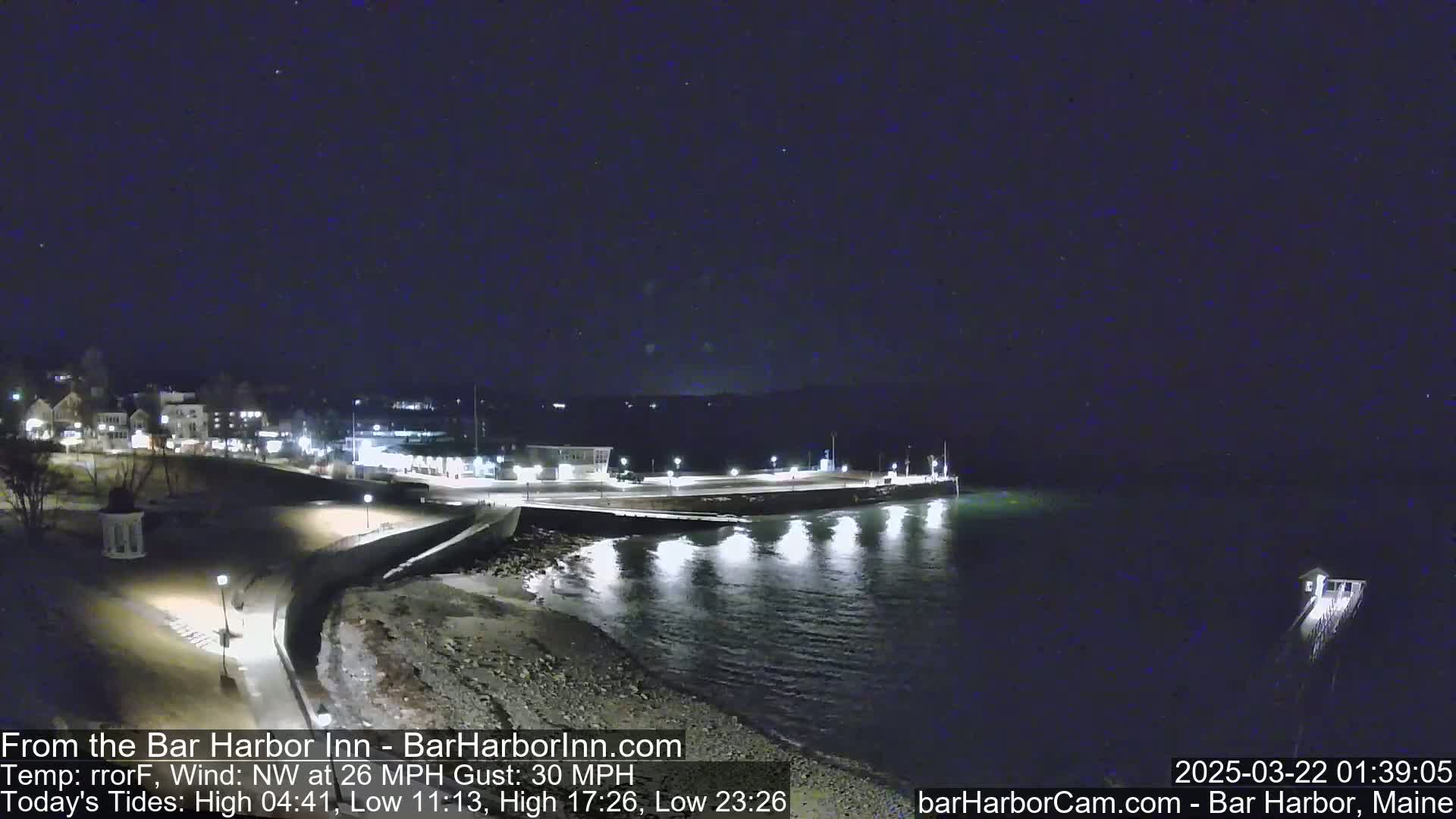 A nighttime coastal scene shows a town's lights along a shoreline with a calm, dark body of water under a clear, starry sky, with a northwest wind at 26 mph and gusts up to 30 mph.