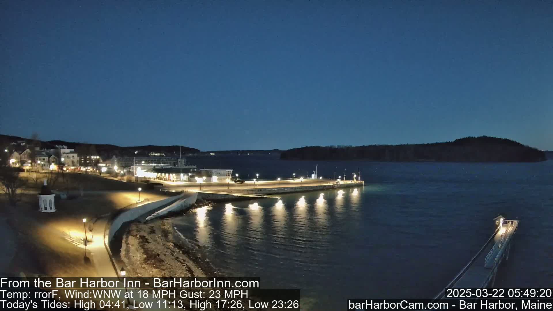 A nighttime view of a harbor town with a long pier extending into calm, dark water under a clear, twilight sky.
