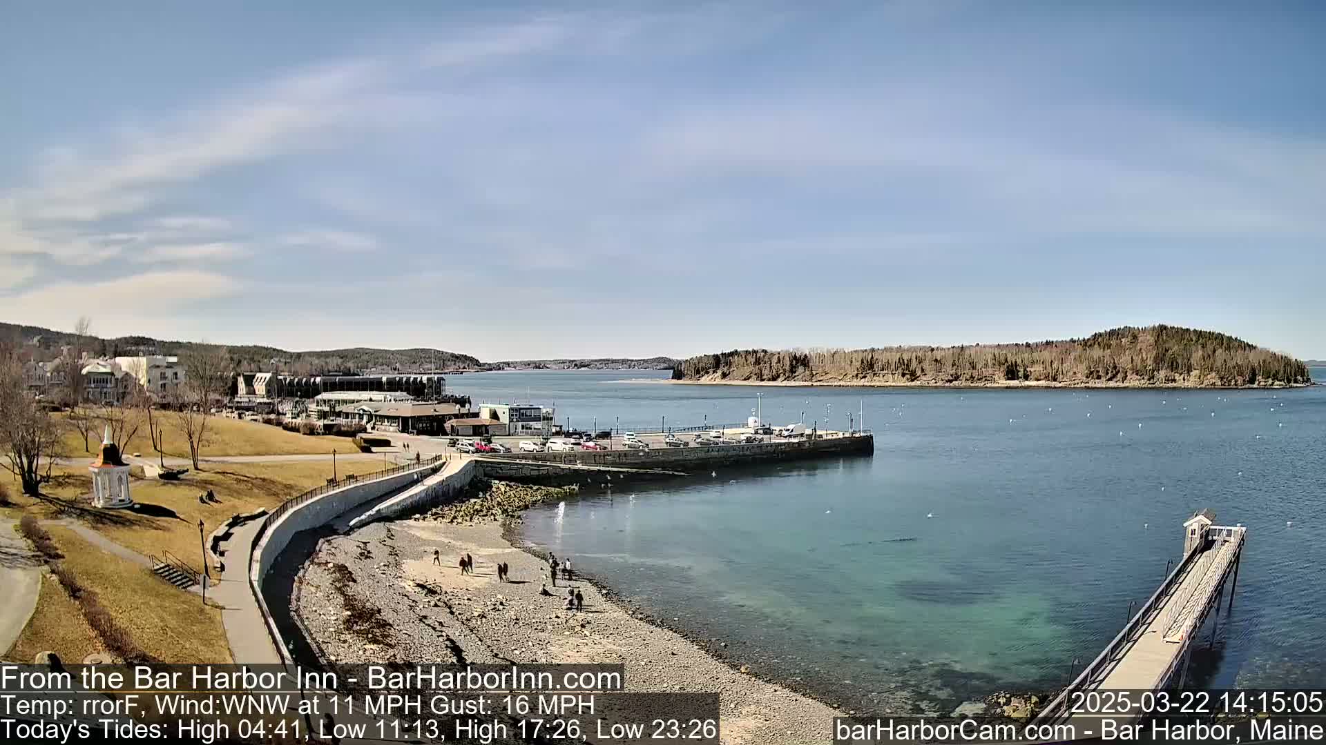 A seaside town with a harbor, a small island, and a long pier is visible on a partly sunny day with a light wind.