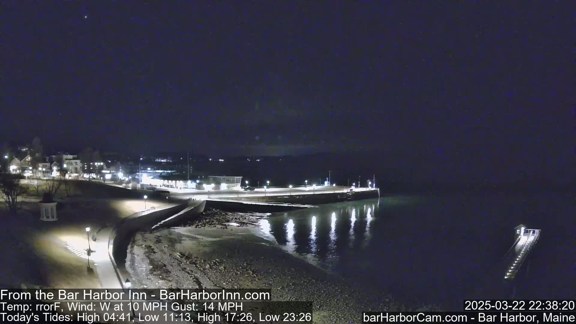 A nighttime coastal scene shows a calm, dark body of water reflecting lights from a nearby town and pier under a clear, dark sky.