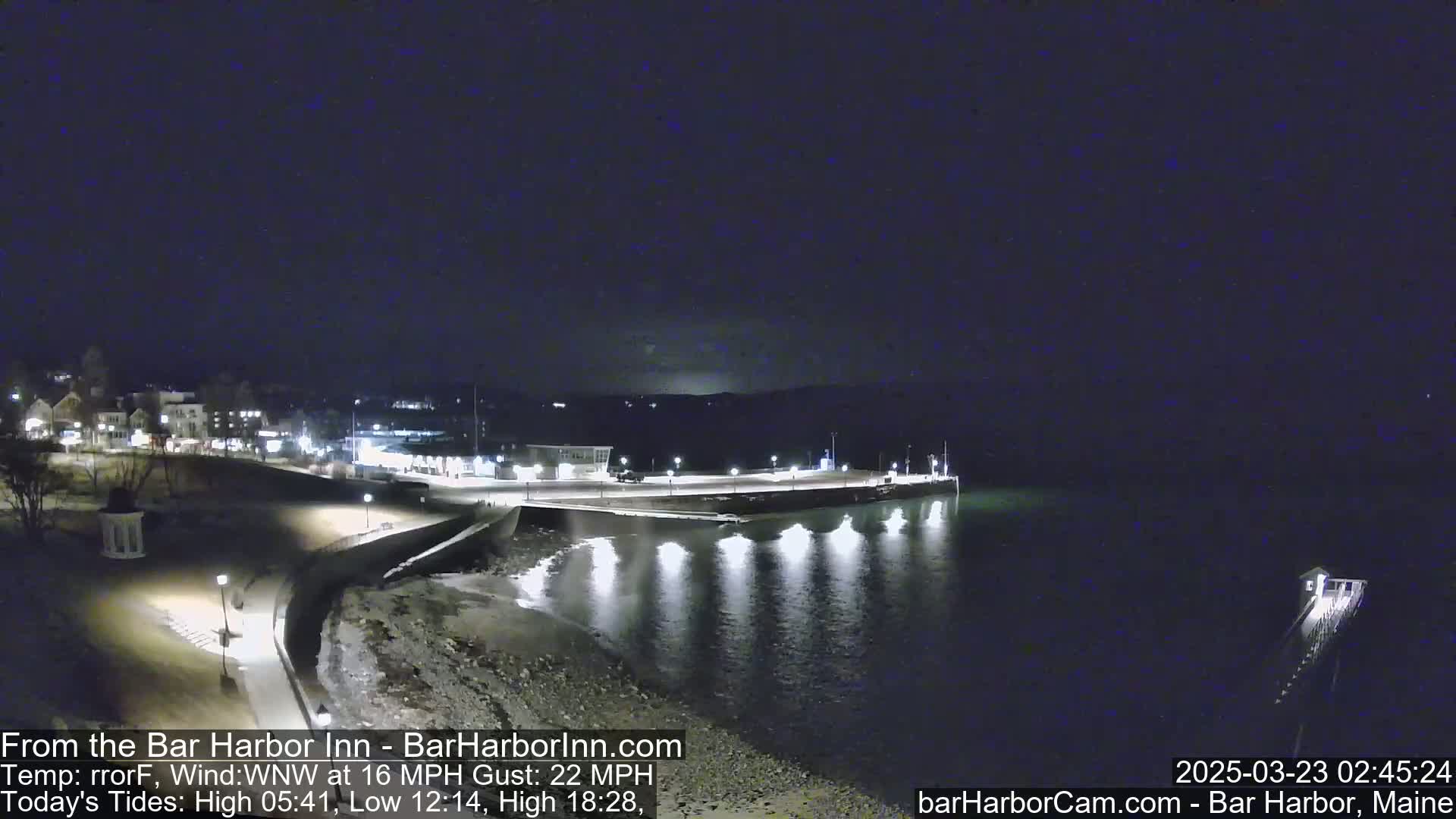 A nighttime view of a harbor with a town in the background, under a clear, dark sky with a 16 mph wind and gusts up to 22 mph.