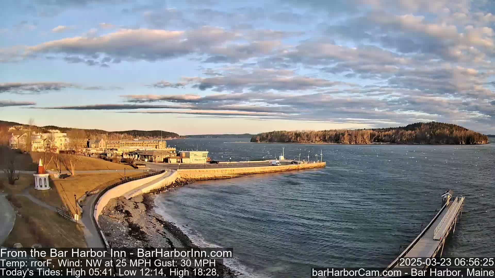 A waterfront scene with a large hotel, a stone seawall, and a long pier extending into a choppy sea under a partly cloudy sky.
