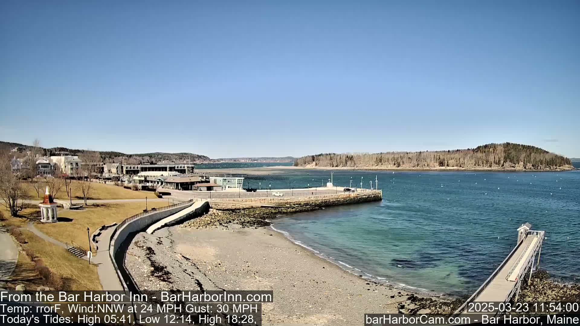 A clear, sunny day shows a rocky beach, a harbor with buildings and a long pier extending into the water, under a bright blue sky.