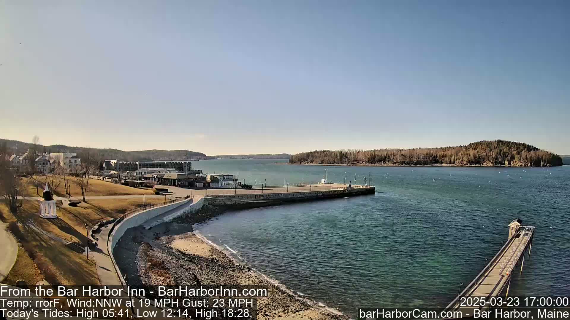 A clear, sunny day shows a coastal town with a long pier extending into calm water, with a small island visible in the distance.