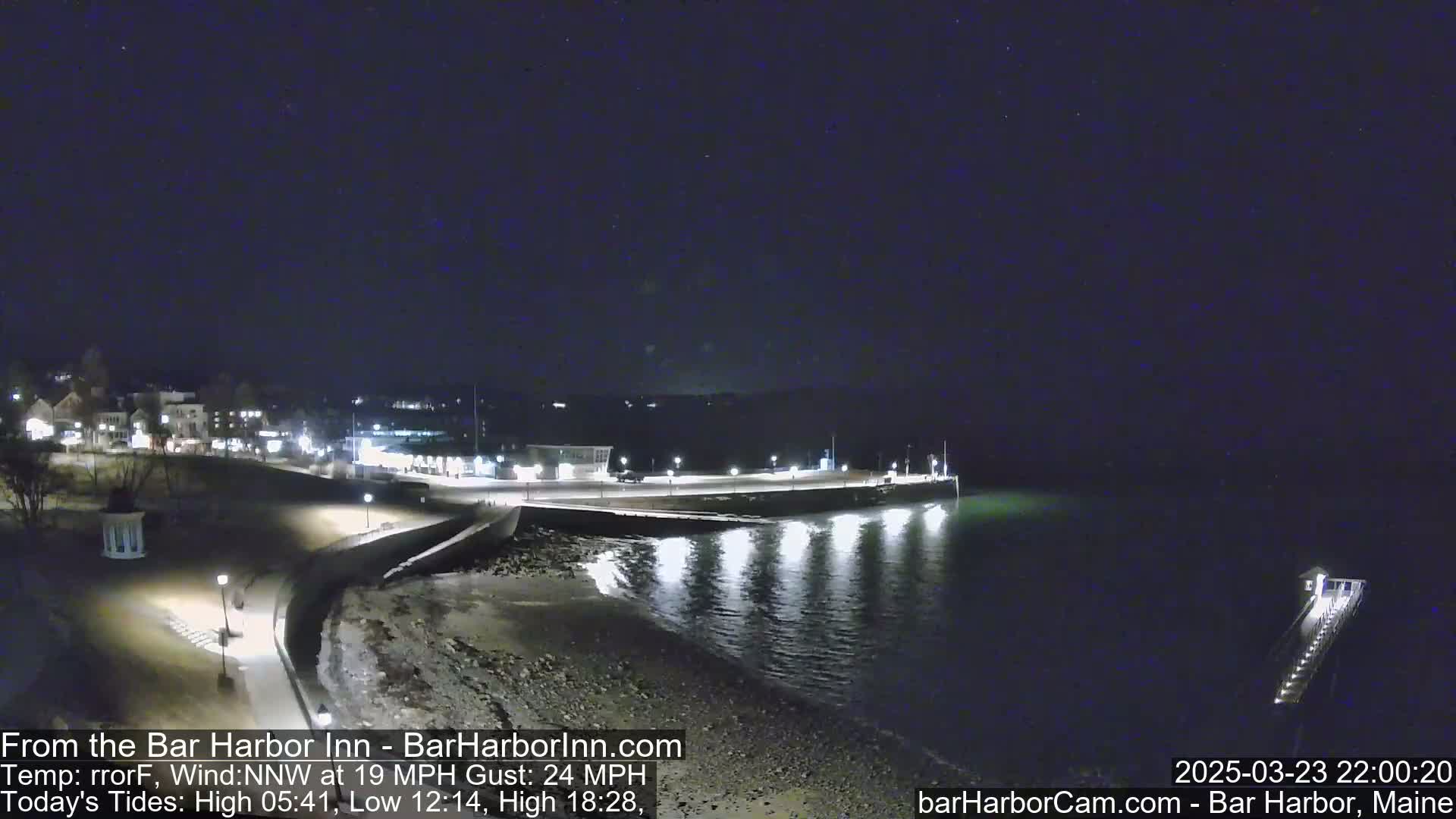 A nighttime view of a coastal town with a calm, dark body of water reflecting lights from a pier and buildings on shore under a clear, dark sky.