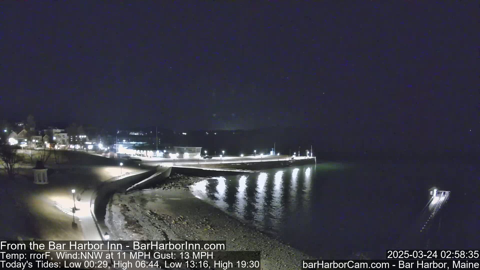 A nighttime view of a calm harbor with a town's lights reflecting on the water under a clear, dark sky.