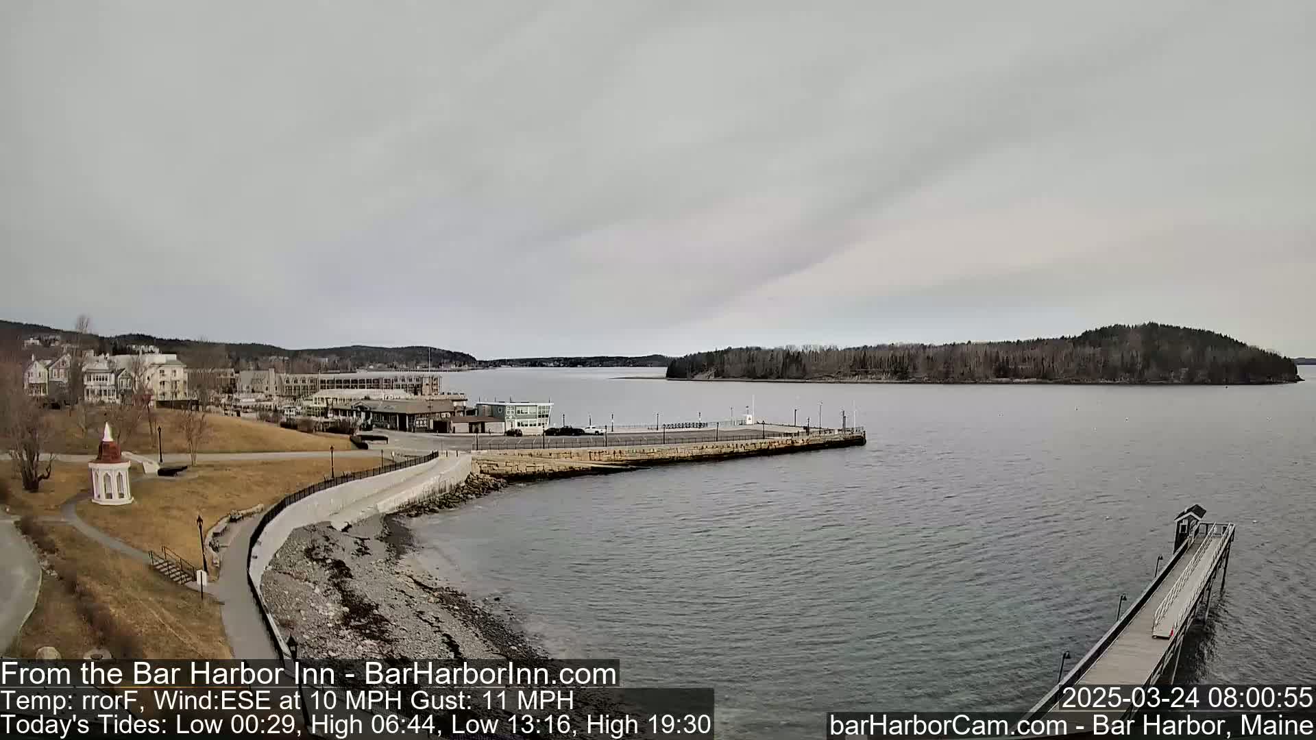 A mostly cloudy day shows a harbor town with buildings, a small gazebo, and a long pier extending into calm gray water, with a small island visible in the distance.