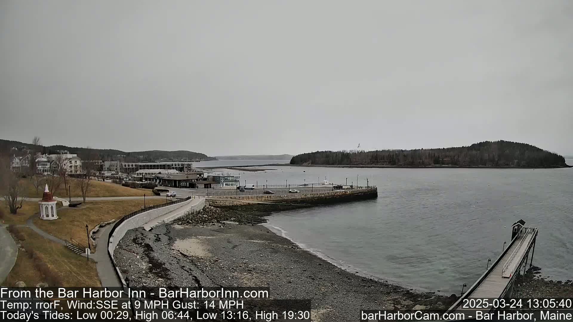 A coastal scene on an overcast day shows a rocky shoreline, a calm harbor with a small island in the distance, and a town visible in the background.